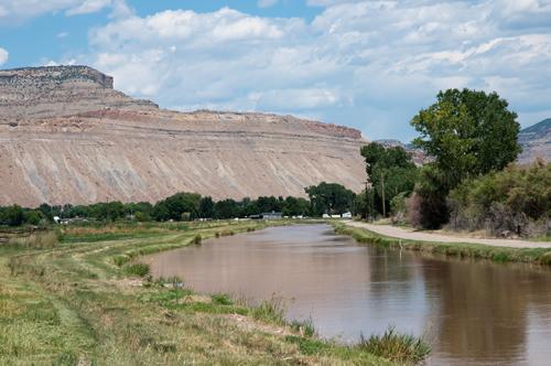 Irrigation ditch full of water with trees growing on the bank and the desert mountains and cliffs that make up the Bookcliffs that stretch almost 200 miles across western Colorado and eastern Utah.