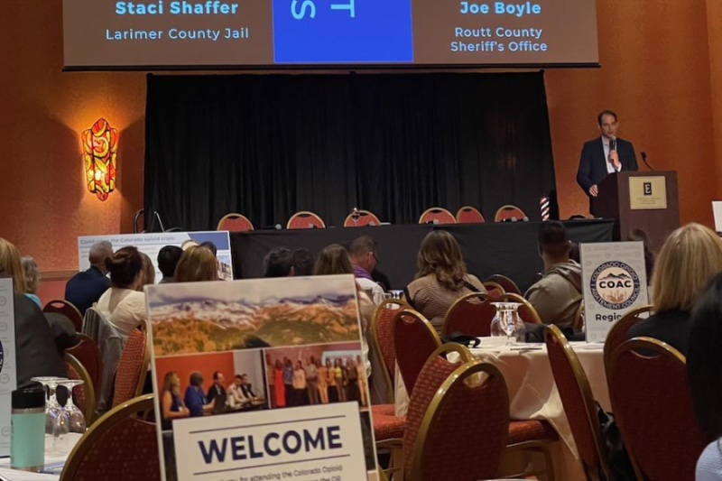 Speaker presenting at the 3rd Annual Opioid Abatement Conference with names of panelists displayed on a large screen, and COAC region signage visible on tables in the foreground.