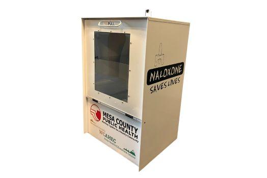 Image of white, metal kiosk with a handle at the top of a glass door. It includes a sticker on the side that reads naloxone saves lives.