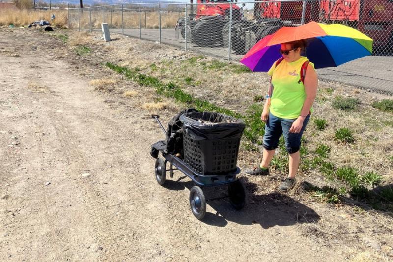 Woman standing under rainbow umbrella under black trash bin on wheels in open space. 