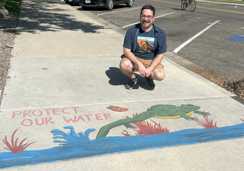 Artist Andrew West kneeling by his stormwater art project Protect Our Water that depicts a green frog leaping up out of a river. 