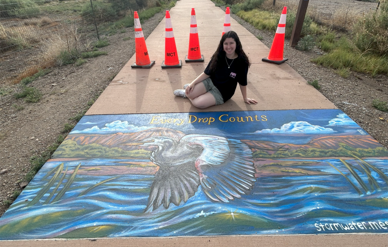 Artist Mindy Hill sitting on the ground in front of her stormwater drain art project that depicts a blue heron flying over the river. 