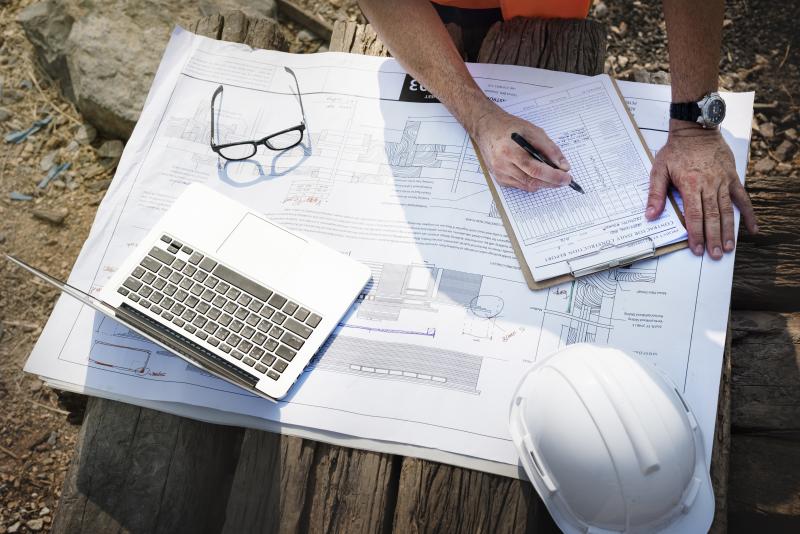 A site engineer with a white hardhat and a laptop is checking off a list on a clipboard, which are sitting on top of his construction plans.