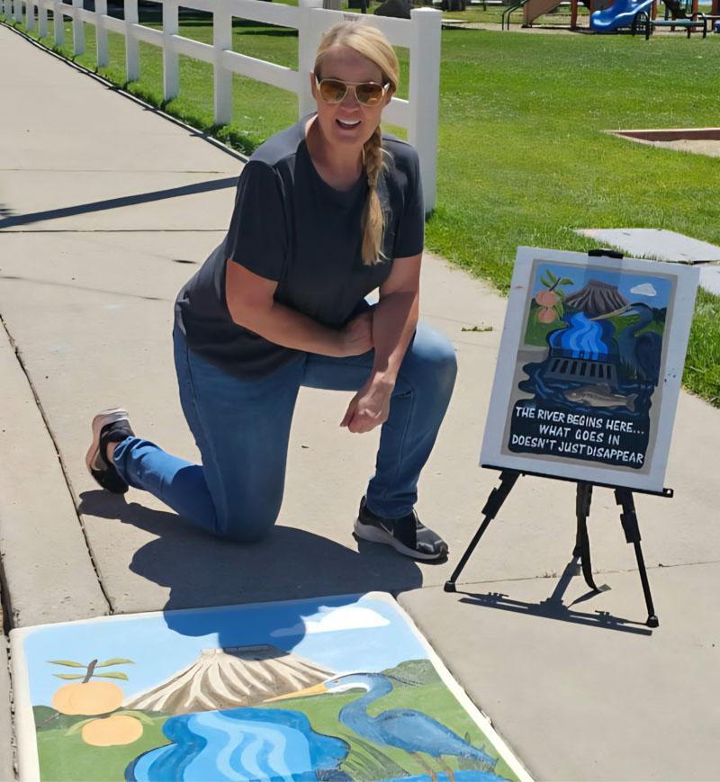 Artist Carolyn Stifflear kneeling next to her stormwater project and a smaller copy of it sitting on an easel; the piece depicts the water run-off from Mount Garfield over the orchards of Palisade.