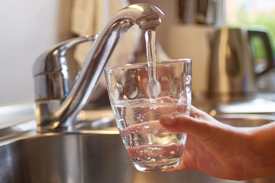 Hand holding glass under running faucet in kitchen. Cup is more than half full of water.