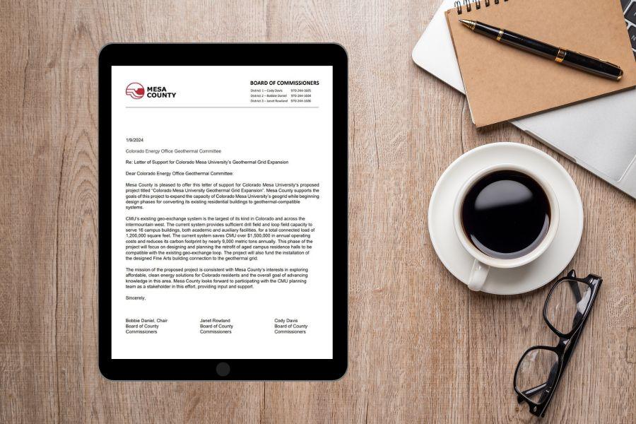 Aerial view of brown wood desk with a brown notebook and black and gold pen next to a cup of black coffee in a white cup and a black iPad displaying Mesa County's letter of support for CMU's geo-thermal expansion. 