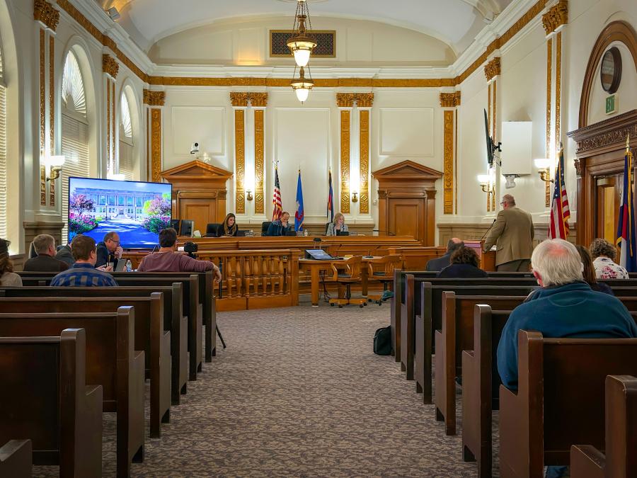 Room full of people sitting in official government room with dark wood benches, white walls, and bright lights. 