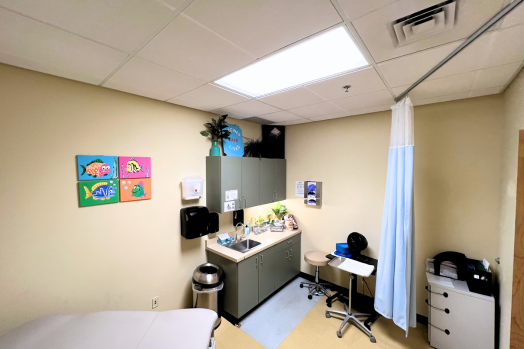 Inside a room in the Public Health Clinic showing a patient table, sink, cupboards, and art on the wall.