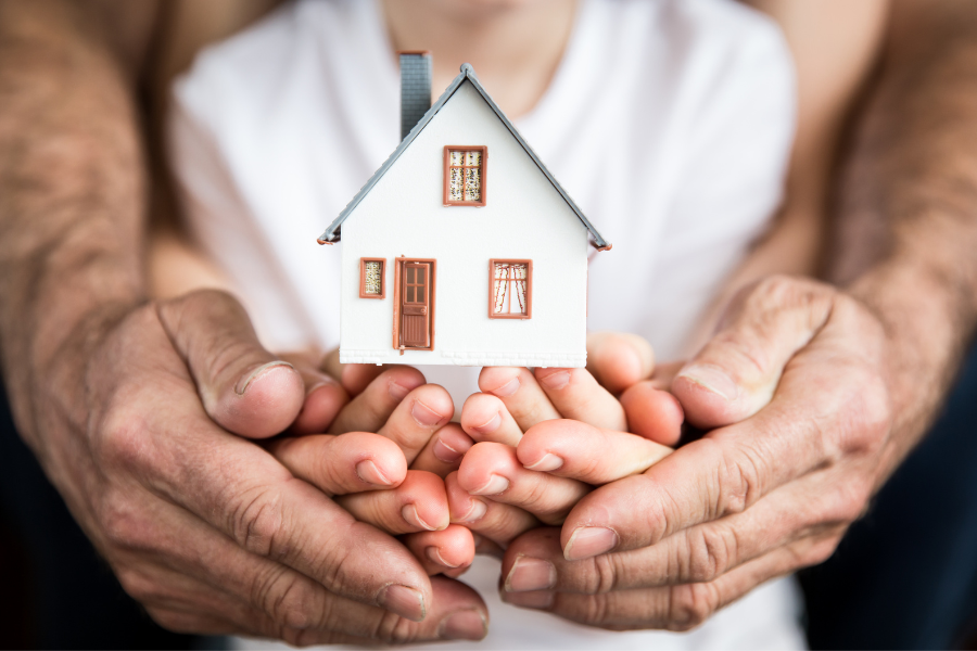 Adult hands hold child hands that hold small white, brown and blue house. 