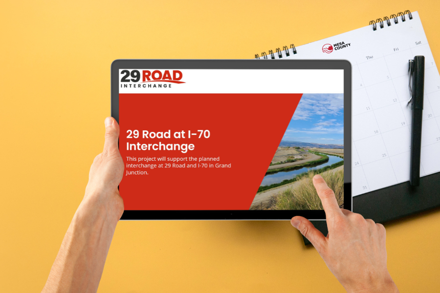 Yellow background with part of a calendar showing and two hands holding an iPad displaying an invite with a red background and white text reading, "29 Road at I-70 Interchange."