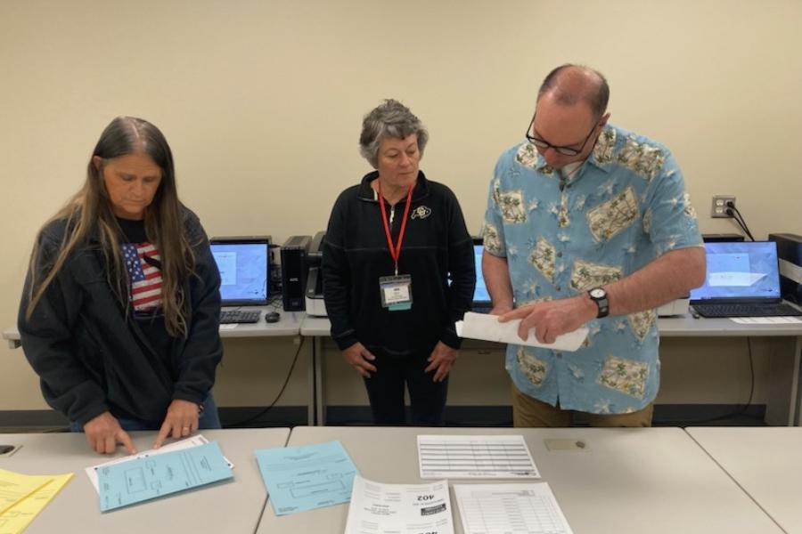 3 people standing above table looking at white and blue papers. 