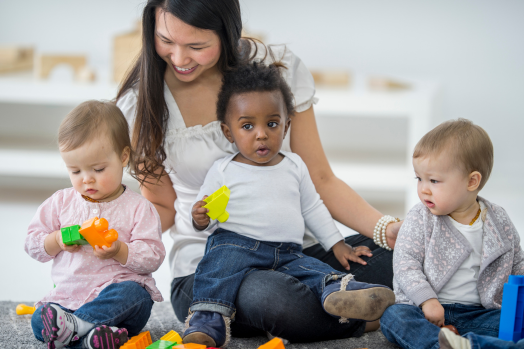 Three young children playing with toys on the ground by their caregiver, who is a woman with long, dark hair.