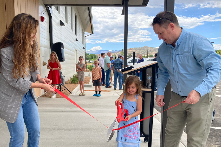 Man and woman holding a red ribbon with a child holding big red scissors. 