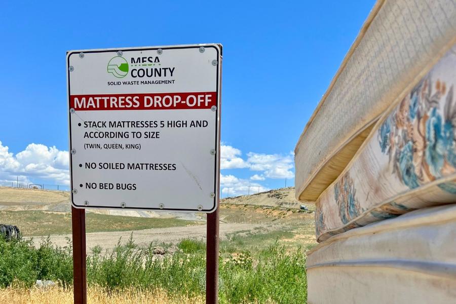 Three stacked mattresses to the right of a sign with black and green Mesa County Solid Waste Management logo with red banner and white text over reading, "MATTRESS DROP-OFF, " and black text reading, "STACK MATTRESSES 5 HIGH AND ACCORDING TO SIZE (TWIN, QUEEN, KING) NO SOILED MATTRESSES NO BED BUGS."