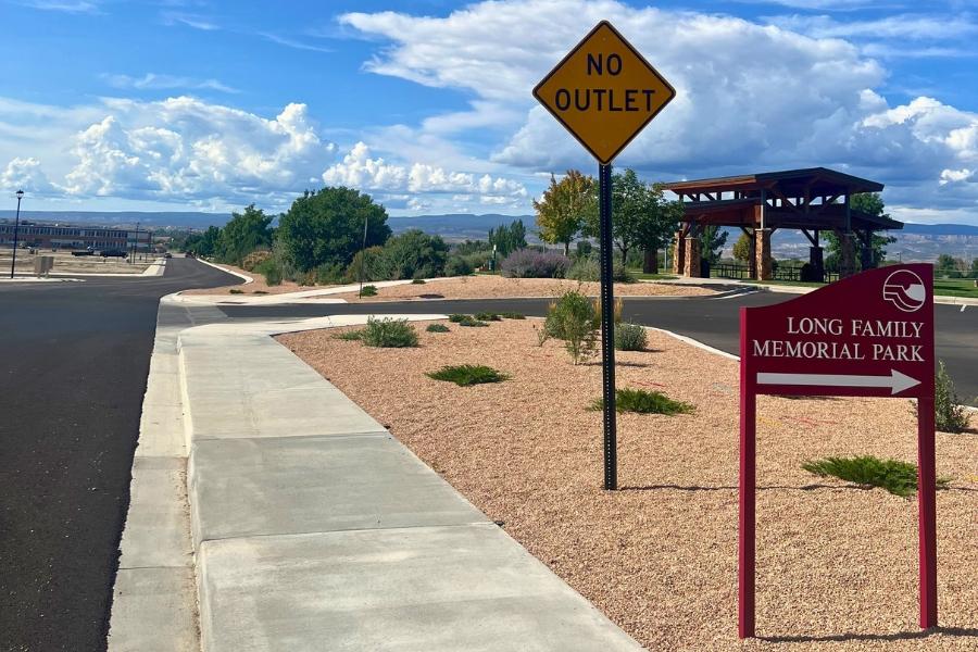 Park entrance in front of blue sky covered in white clouds with red sign reading long family memorial park in white text with an arrow pointing right and the mesa county logo next to a yellow sign reading no outlet in black text.