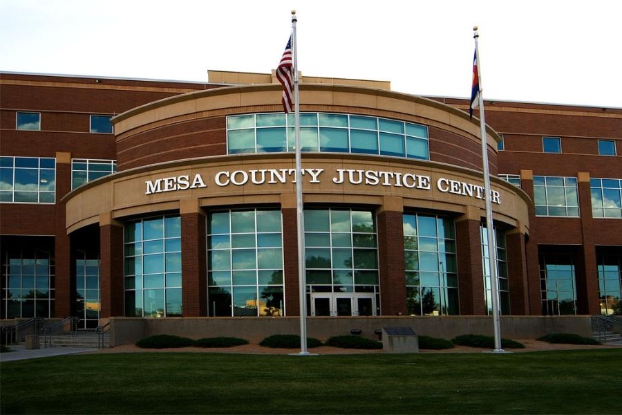 Brick building with white letters reading mesa county justice center and an American flag and Colorado flag in the green grass in front of the building. 