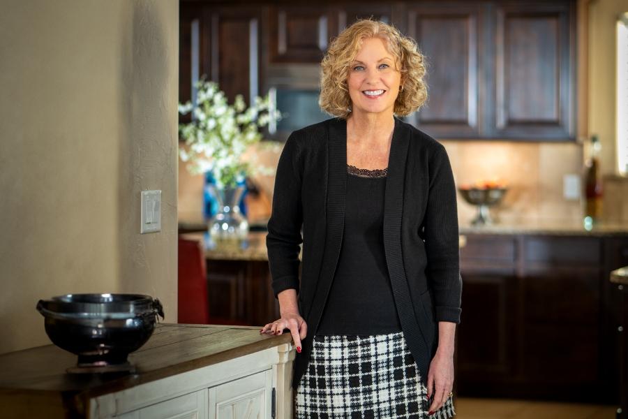 Woman (Janet Rowland) smiling in kitchen with hand sitting on counter. 