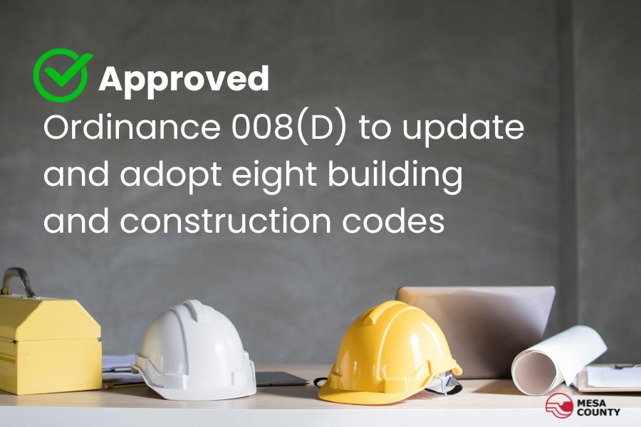 White and yellow hard hats sitting on a desk with white text above reading, Approved Ordinance 008(D) to update and adopt eight building and construction codes with the red and black Mesa County logo. 