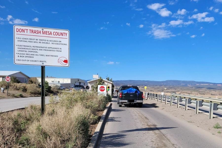 A black truck with a blue tarp covering the truck bed enters the Mesa County Landfill with a white sign on the left with red text reading, don't trash Mesa County. 