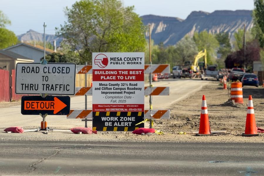 Road construction sign that reads, road closed to thru traffic, with an orange detour sign pointing right, next to a Mesa County road project sign. 