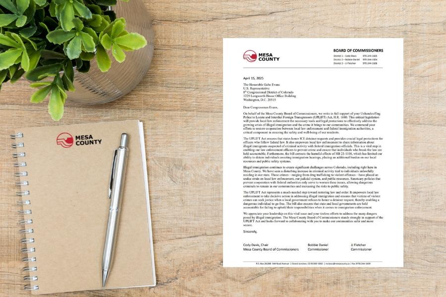 A letter sitting on a wood table next to a tan notebook with the Mesa County logo on it and a house plant. 