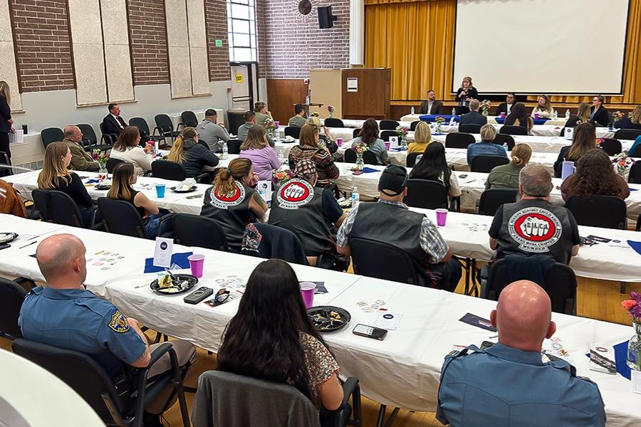 A large group of people sits at decorated tables in a gymnasium-style room for a formal event. Attendees include uniformed law enforcement officers, community members, and individuals wearing vests with the Bikers Against Child Abuse (BACA) logo. 