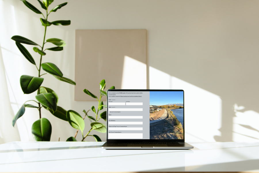 A laptop on a white desk in a bright room with large green plants. The screen displays a public comment submission form for the Stormwater Management Manual, featuring fields for contact information and a section to submit feedback.