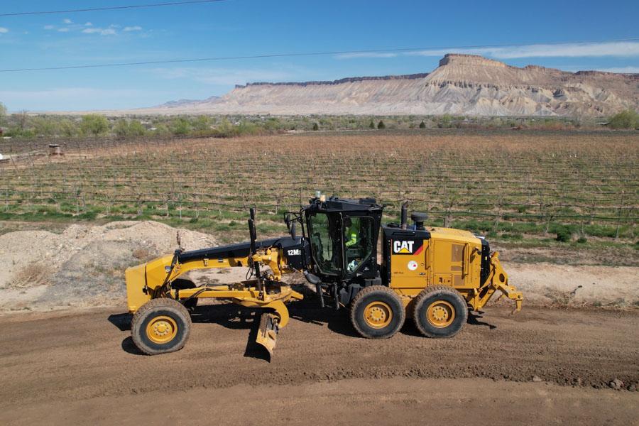 A yellow Caterpillar motor grader works on a dirt road in a rural area with vineyards in the background. The operator is visible inside the cab, wearing safety gear. The scenic backdrop includes Mount Garfield under a clear blue sky.