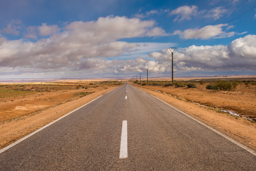A straight, two-lane road stretching into the distance through a wide, open desert landscape, with power poles lining the roadside and dramatic clouds filling the sky.
