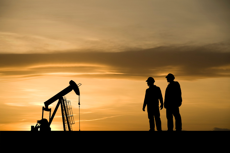 Silhouetted oil pumpjack and two workers wearing hard hats stand on a ridgeline at sunset, with an orange and gold sky in the background.