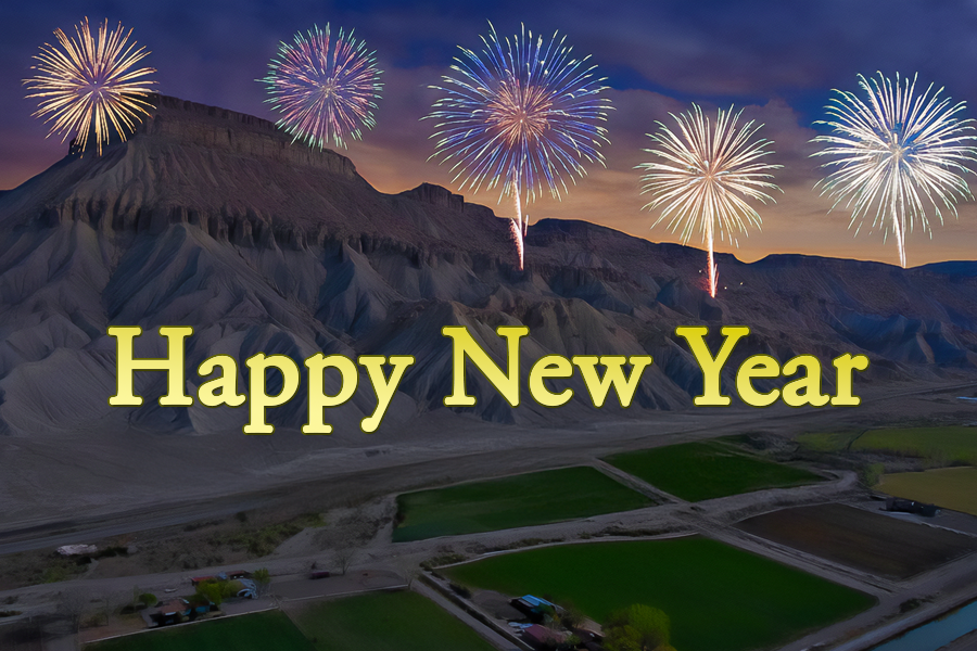 Fireworks burst in the night sky above Mt. Garfield and a valley of fields, with large yellow text in the foreground reading “Happy New Year.”