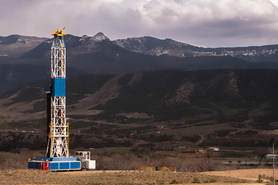 Oil and gas drilling rig in a rural landscape, with foothills and mountain ridgelines in the background.