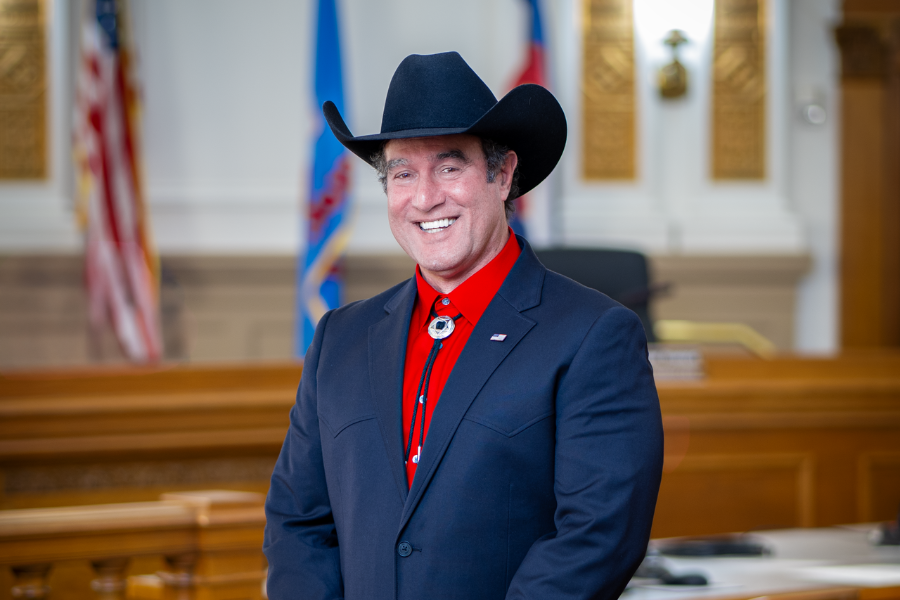 A smiling JJ Fletcher wearing a black cowboy hat, dark suit and red shirt stands in the public hearing room, with flags and wood-paneled desks visible in the background.
