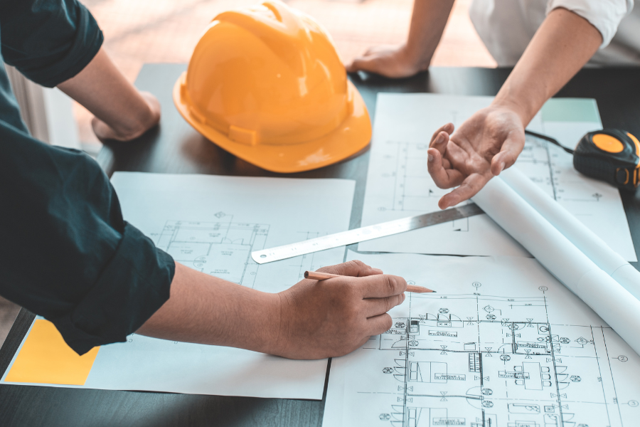 Two people review architectural or engineering plans spread out on a table, with a yellow hard hat, ruler and rolled blueprints nearby.