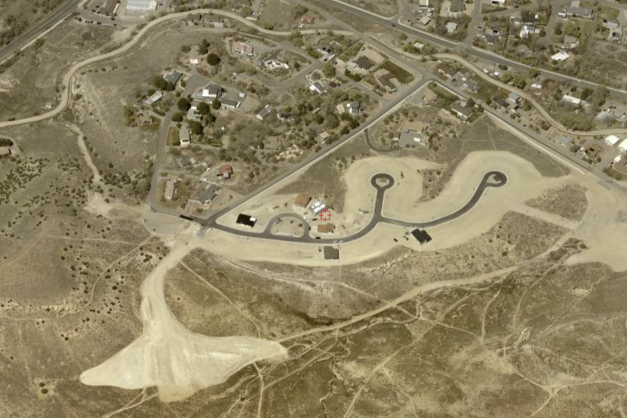 An aerial view of a developing residential area with newly paved roads forming cul-de-sacs, surrounded by open desert terrain and nearby established neighborhoods. A red marker highlights one property within the new development.