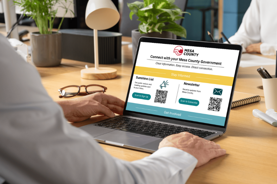Person using a laptop displaying a Mesa County webpage titled “Connect with Your Mesa County Government,” featuring options to sign up for updates, volunteer, report road issues and explore online resources.