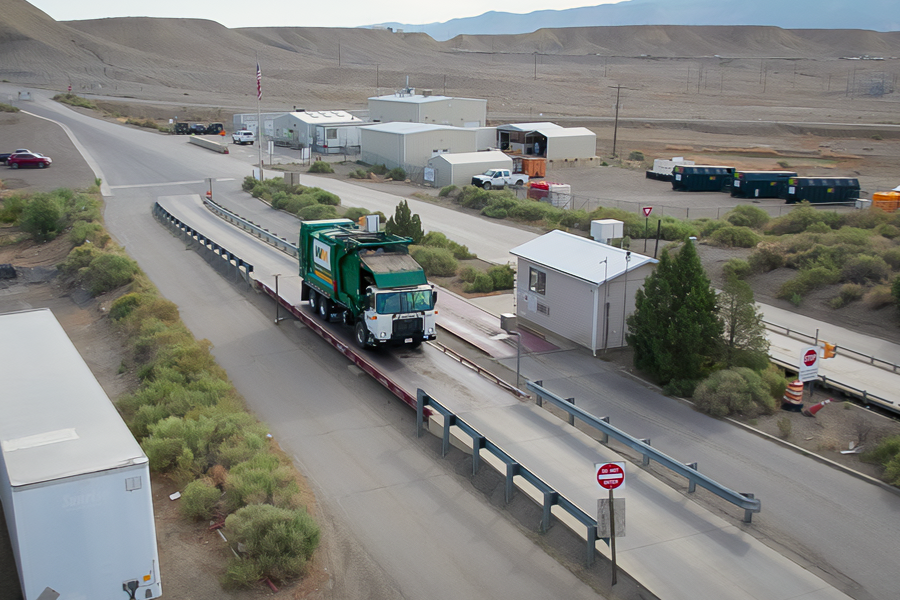 Mesa County landfill entrance with a garbage truck driving across a scale, surrounded by small buildings, equipment containers and desert hills in the background.