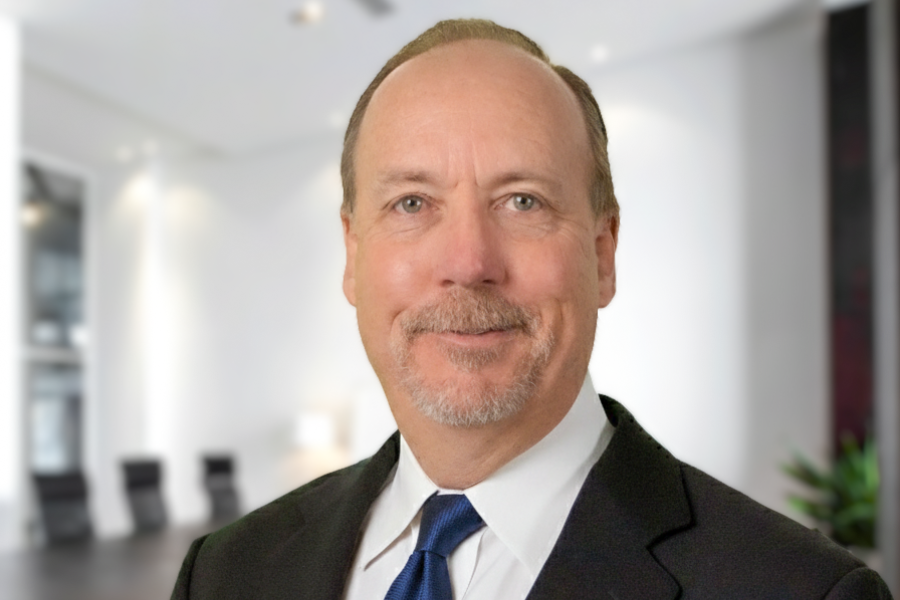 Portrait of Mesa County Attorney Todd Starr wearing a dark suit, white shirt and blue tie, standing against a softly blurred office background.