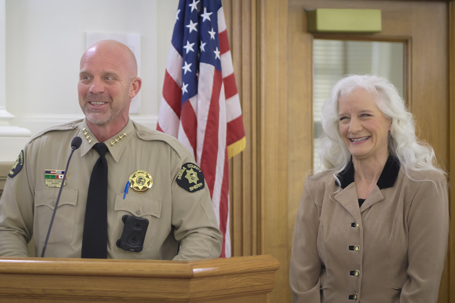 Mesa County Sheriff Todd Rowell speaks at a podium during a public hearing as Laurie Galvin stands beside him smiling, with an American flag in the background.