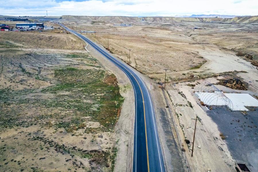 An aerial view of a newly paved rural road curving through an open desert landscape, with scattered buildings, utility poles and distant hills under a cloudy sky.
