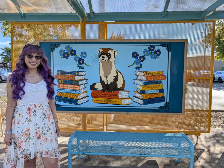 Muralist Mae Pina standing in front of her mural of a black footed ferret sitting on a stack of books with books to the right and left