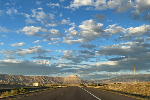 A highway stretches towards a Mt. Garfield and the Grand Mesa under a vast sky filled with scattered white and gray clouds.