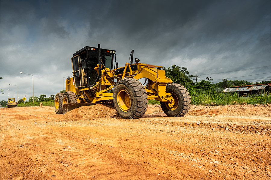 Motor grader spreading and leveling gravel on an unpaved road under dark storm clouds during roadway maintenance work.