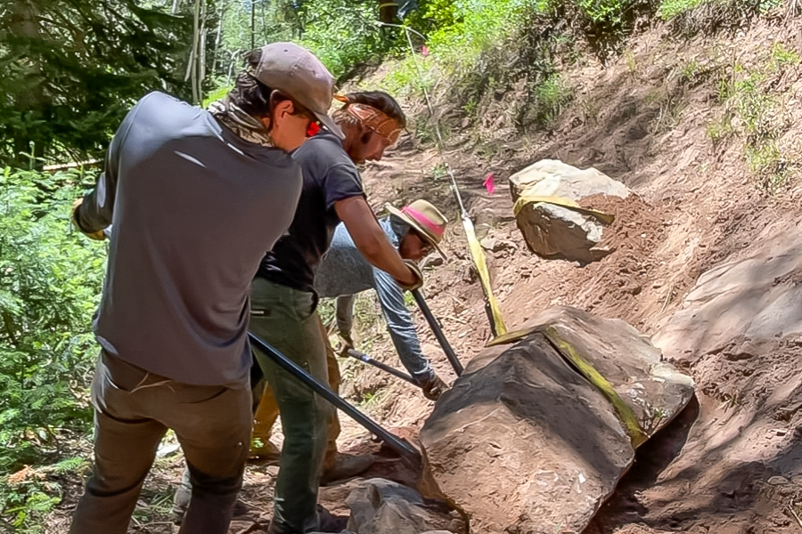 Trail crew members use tools to move large rocks and repair a dirt trail on a forested hillside in the Turner Gulch burn area.