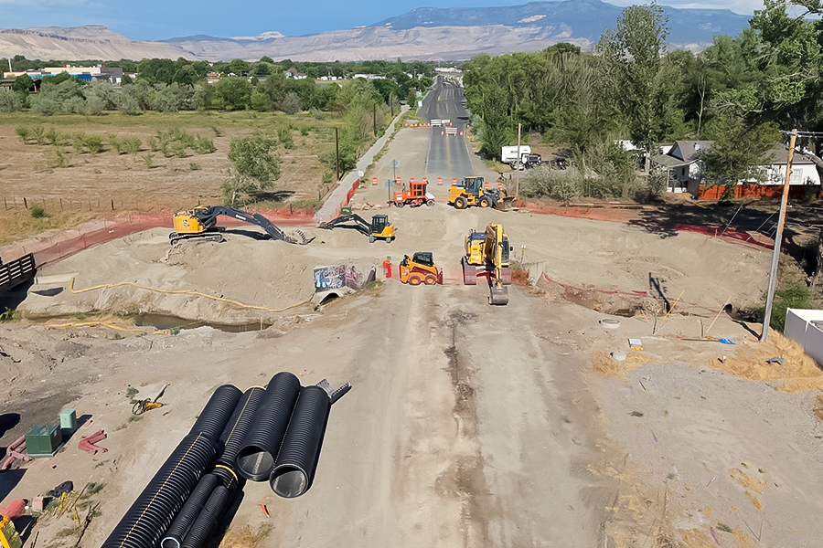 An aerial view of active road construction on Orchard Avenue shows excavators, loaders and drainage pipe installation, with traffic control in place and the Grand Mesa visible in the background. 